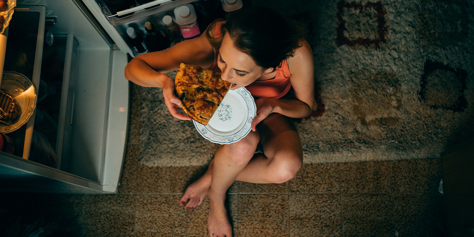 woman eating pizza by an open fridge